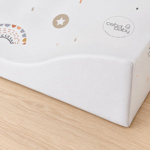 Close-up of the Sleeping Bear & Balloons baby changing mat showing its curved padded corner, white textured surface and rainbow and star details on a light oak dresser.