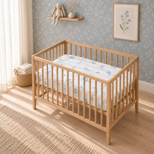 White baby cot fitted sheet with small blue forget-me-not flowers on a light oak cot in a softly styled nursery. Fabricco UK