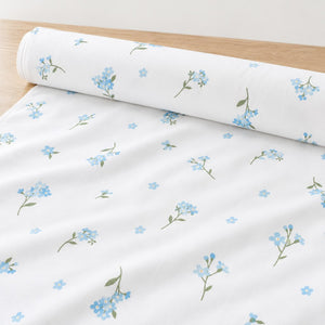 Roll of Forget-me-not cotton fabric with pale blue flowers and green stems on white, unrolled on a light oak table