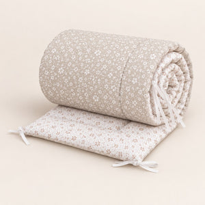 Padded meadow floral cot bumper lying partly unrolled, showing the light beige floral outer, beige-on-white reverse and narrow white ties.