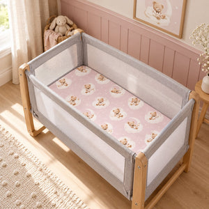 Pink teddy bear and clouds fitted sheet on a bedside crib mattress in a soft pink baby girl’s nursery.