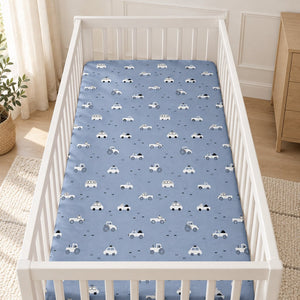 Blue cars and tractors cotton fitted sheet on a white cot mattress in a Scandinavian nursery with light wood furniture.