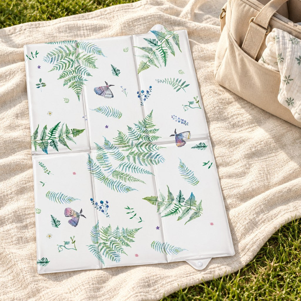 Botanical and butterfly baby changing mat open on a cream picnic blanket on grass, beside a beige changing bag.