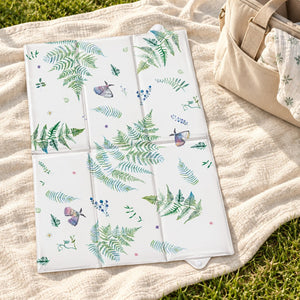 Botanical and butterfly baby changing mat open on a cream picnic blanket on grass, beside a beige changing bag.