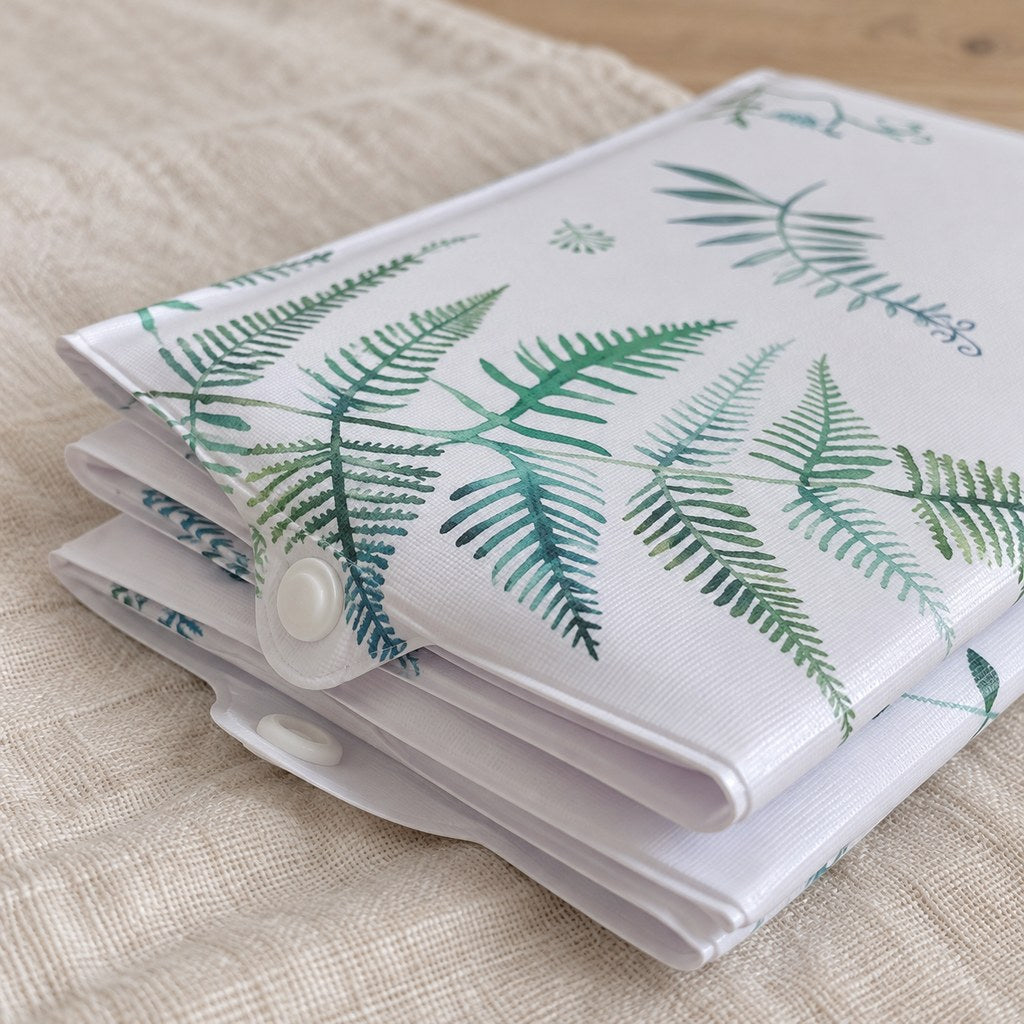 Close-up of the botanical travel changing mat’s white popper tab, green leaf print and thin folded layers on cream linen.