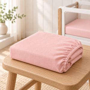 Folded blush pink SnuzPod fitted sheet in 100% cotton beside a bedside crib