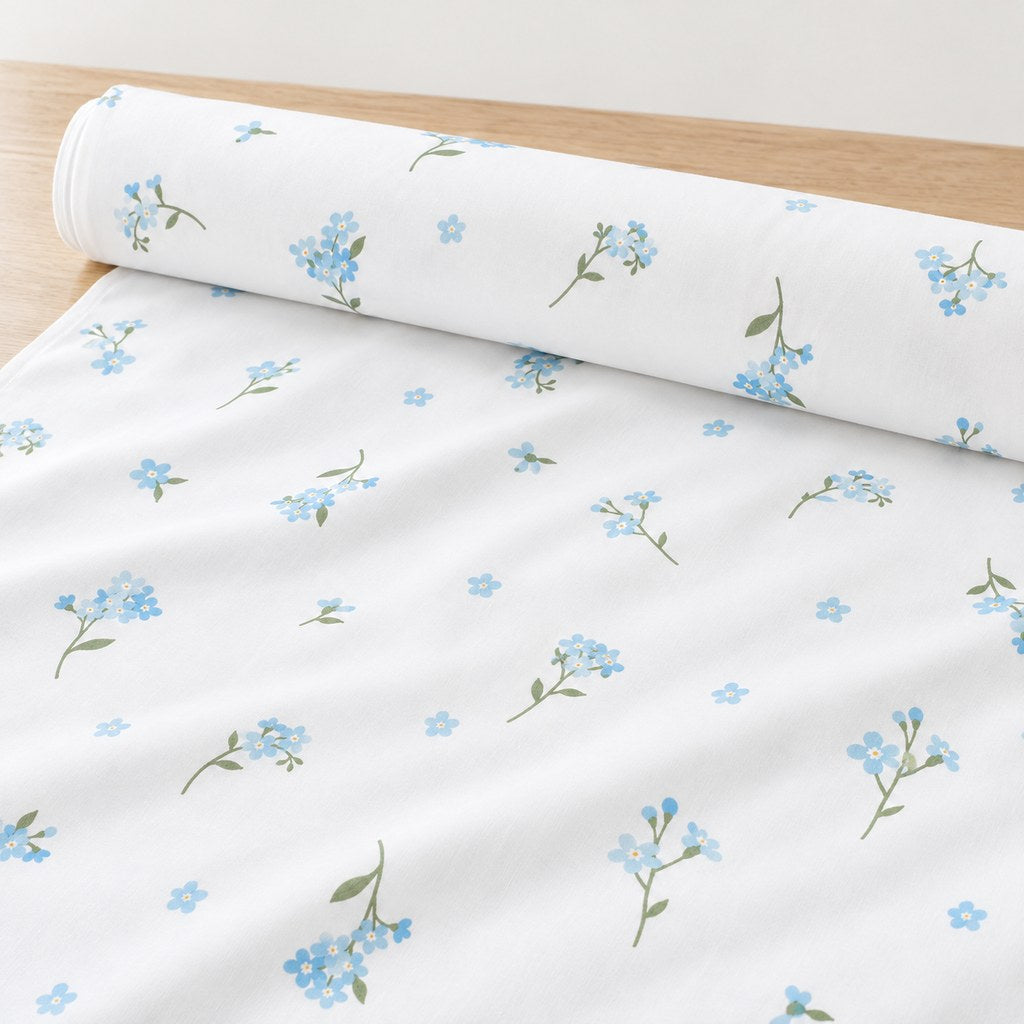 Roll of Forget-me-not cotton fabric with pale blue flowers and green stems on white, unrolled on a light oak table