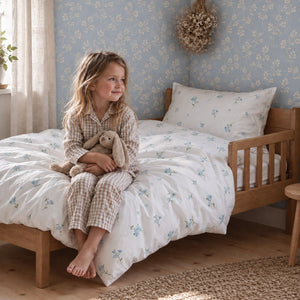 Child sitting on an oak cot bed dressed in white Forget Me Not floral bedding, with a pale blue botanical wall behind. Fabricco UK