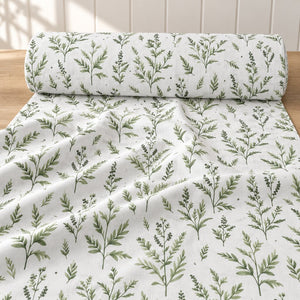 Roll of white cotton fabric with green leafy branches, partly unrolled on a light wood table