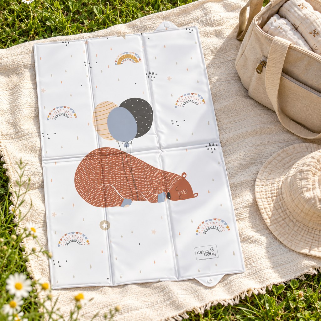Sleeping bear and balloons baby changing mat open on a cream picnic blanket on grass, beside a beige changing bag and sunhat.