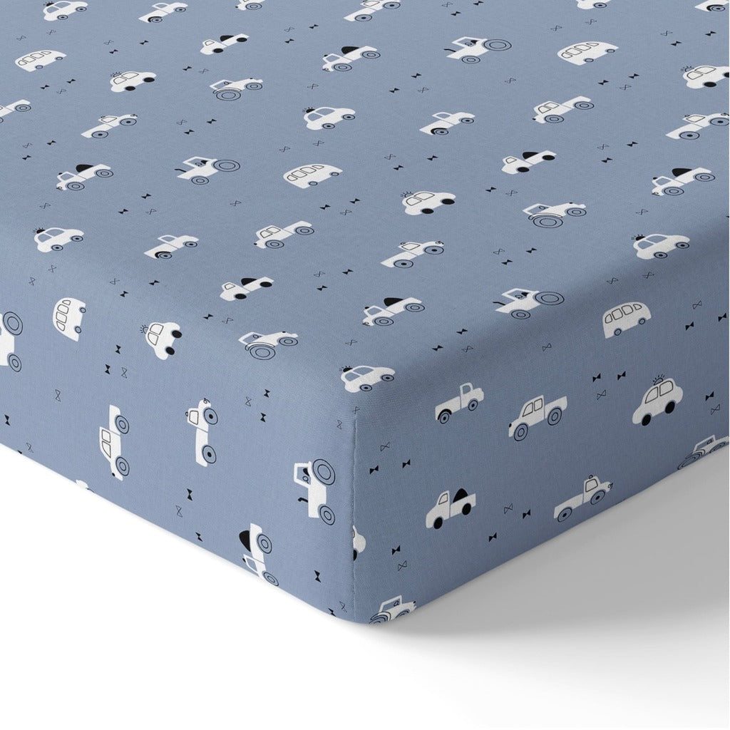 Close-up of a blue cotton baby fitted sheet fitted around a mattress corner, showing white cars, tractors, buses and small black bow details.