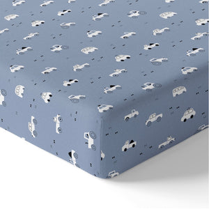 Close-up of a blue cotton baby fitted sheet fitted around a mattress corner, showing white cars, tractors, buses and small black bow details.