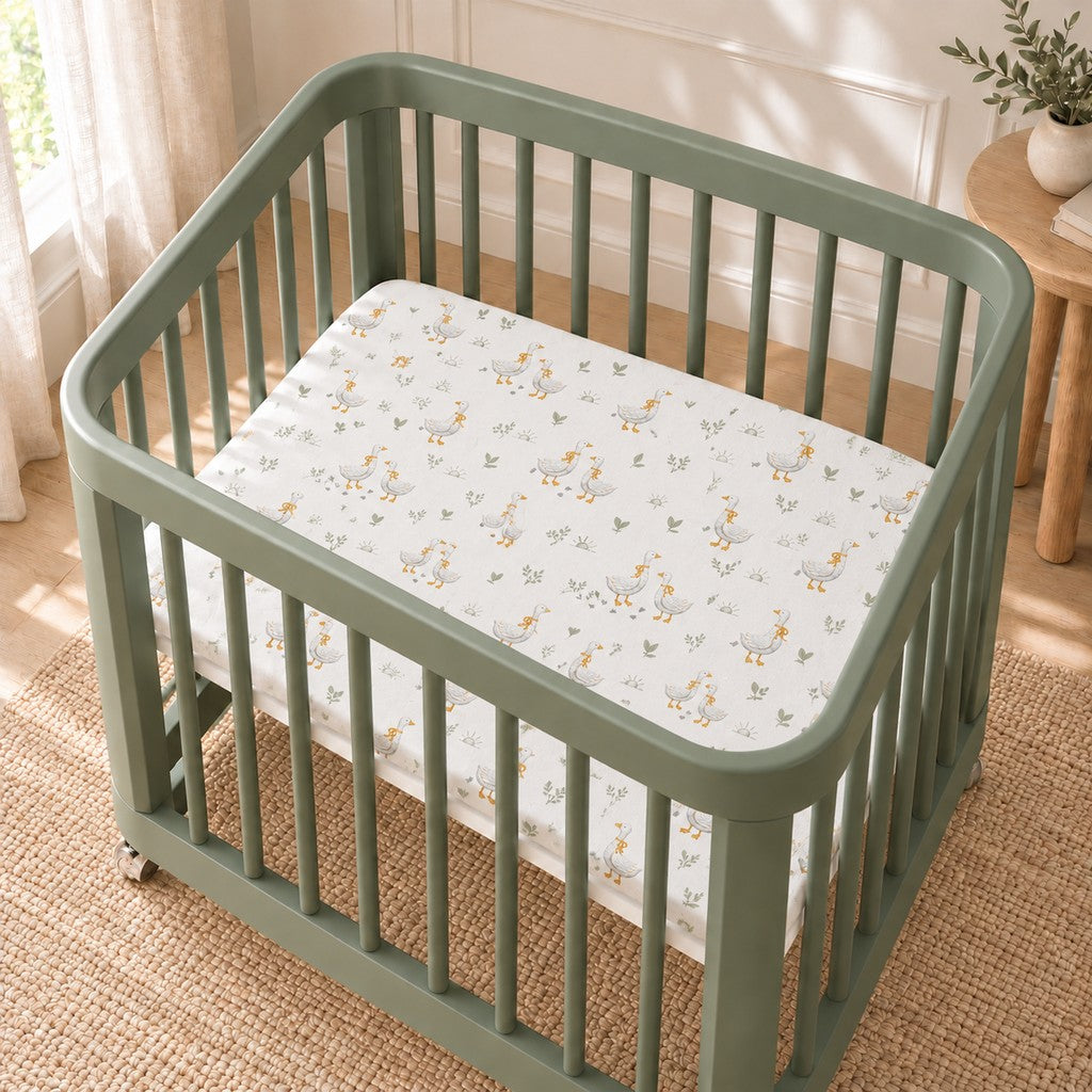 Little Goose fitted sheet with grey geese, mustard bows and sage green leaves on a white background, shown in a sage green Nestig-style mini crib.