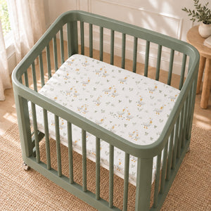 Little Goose fitted sheet with grey geese, mustard bows and sage green leaves on a white background, shown in a sage green Nestig-style mini crib.