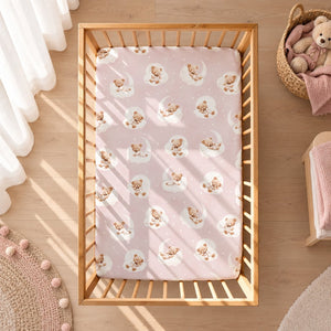 Pink teddy bear and clouds fitted sheet on a wooden cot mattress, featuring white moons and stars.
